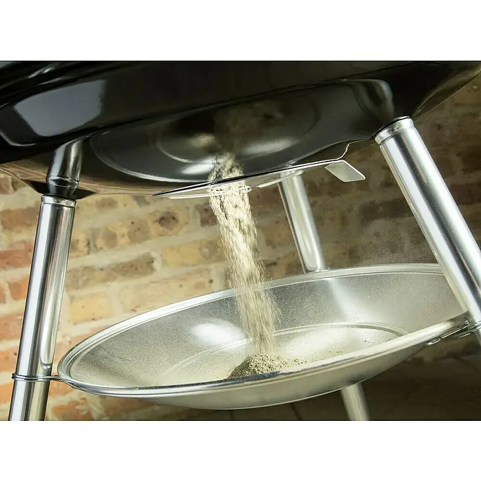 Weber Barbacoa redonda con tapa Compact Kettle - Diámetro de la superficie de la parrilla: 47 cm, Negro - Primer plano de una parrilla esférica mientras se vacían las cenizas del recipiente en un recogedor de cenizas.