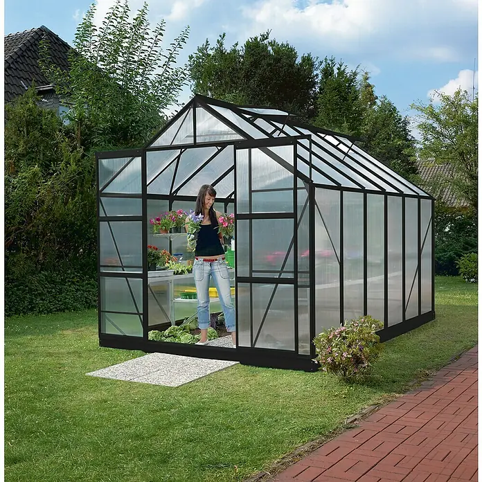Vitavia Gewächshaus Mars 9900 - Außenmaß inkl. Dachüberstand (B x T): 257 x 383 cm, 6 mm, Schwarz - Frau im Gewächshaus mit Blumen. Das Gewächshaus hat eine schwarze Rahmenkonstruktion und eine transparente Verglasung.