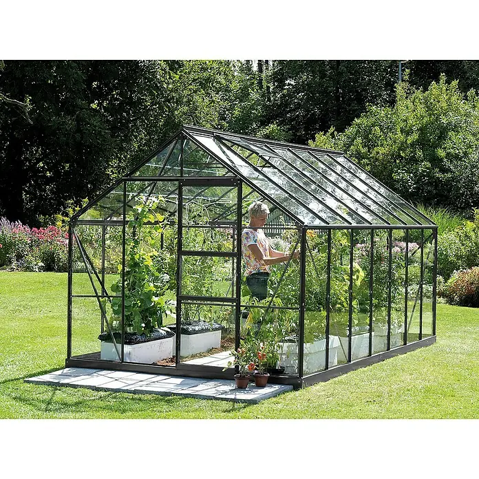 Vitavia Gewächshaus Merkur 11500 Plus - Außenmaß inkl. Dachüberstand (B x T): 257 x 445 cm, 3 mm, Schwarz - Ein großes Gewächshaus aus Glas und Metall steht auf einem Rasen, in dem Gemüse gepflanzt wird. Eine Frau kümmert sich um ihre Pflanzen.