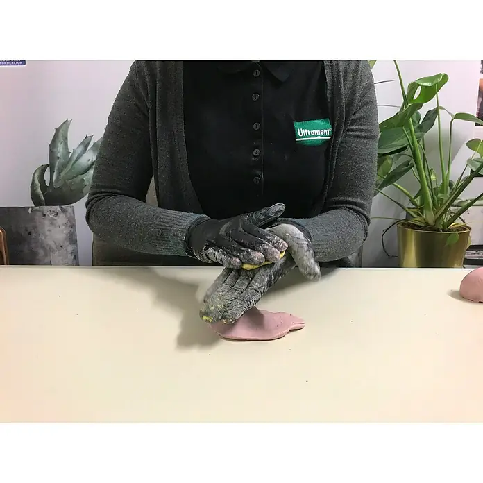 Ultrament Beton-Knetmasse Knet Beton Weiss - Weiss, 2,5 kg - Hände mit schwarzen Handschuhen vermischen gelbe und rosafarbene Modelliermasse auf einem Tisch, während eine Person ein graues Oberteil mit einem Ultrament-Logo trägt.