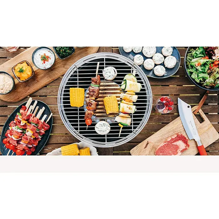 LotusGrill Holzkohlegrill Classic - Durchmesser Grillfläche: 34 cm, Schwarz - Ein runder Grill steht auf einem Holztisch, darauf Spieße, Maiskolben und gefüllte Zucchini, umgeben von Dips, Salat und einem Messer.