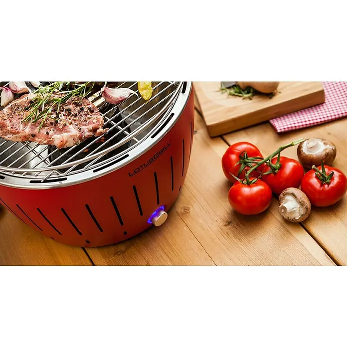 LotusGrill Holzkohlegrill Classic - Durchmesser Grillfläche: 34 cm, Feuerrot - Roter Lotusgrill mit Grillgut, Knoblauch, Rosmarin, Tomaten und Pilzen auf einem Holztisch. Der Lotusgrill ist bereit für ein köstliches Barbecue.