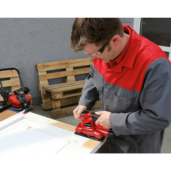 Einhell Schleifpapier-Set - 15 -tlg. - Ein Mann schleift mit einem roten Einhell Exzenterschleifer eine weiße Holztür, um sie für die weitere Bearbeitung vorzubereiten.