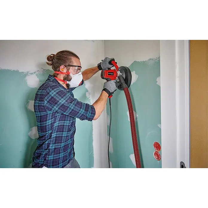 Einhell Schleifgitter-Set - Durchmesser: 180 mm, 5 -tlg., Passend für: Einhell Trockenbauschleifer TE-DW 180 - Ein Mann schleift eine Wand mit einem roten Einhell Trockenbauschleifer, der an einen Staubsauger angeschlossen ist. Er trägt Maske und Schutzbrille.