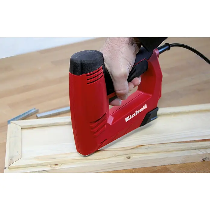 Einhell Feindrahtklammer - Rückenbreite: 11,4 mm, 1.000 Stk. - Eine rote Einhell Elektrotackermaschine wird von einer Hand bedient, um ein Holzstück zu befestigen.