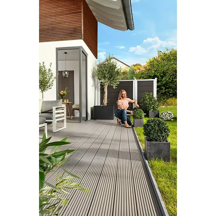Kovalex WPC-Terrassenpaket Exklusiv - Grau, 3 x 3 m - Eine Frau arrangiert Pflanzen auf einer Terrasse mit grauem Holzboden und stilvollen Gartenmöbeln. Die Terrasse bietet einen gemütlichen Außenbereich.