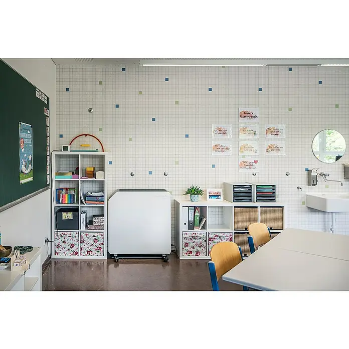 Hönle Luftreiniger SteriWhite Air Q60031,5 x 86 x 86 cm, Mit Standfuß, Max. Luftleistung: 600 m³/h - Classroom-Szene mit Fokus auf ein mobiles Luftreinigungsgerät neben weißen Regalen, die mit bunten Gegenständen gefüllt sind.