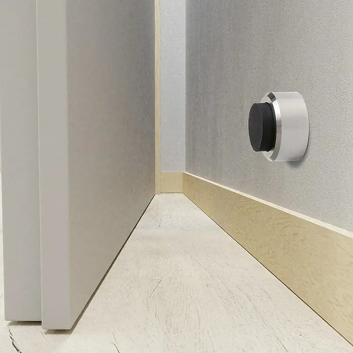 Wandtürstopper - Ø x H: 32 x 15 mm - Nahaufnahme eines modernen Türstoppers mit silbernem Gehäuse und schwarzem Gummipuffer, montiert an einer grauen Wand neben einer Tür.