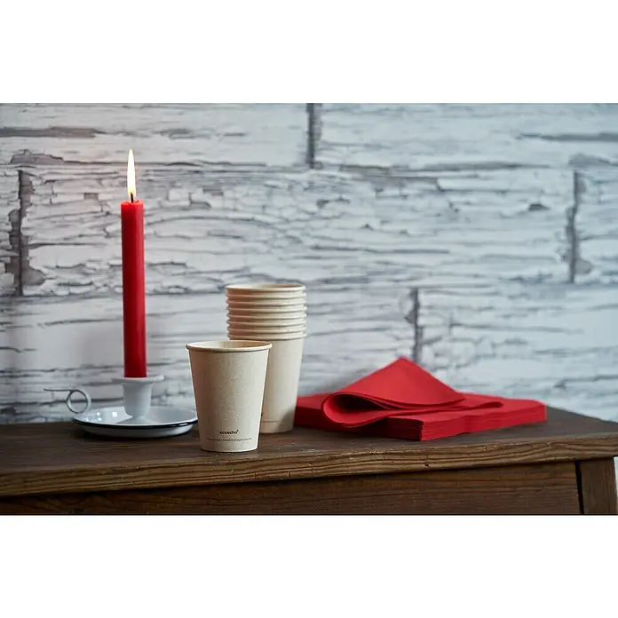 Duni Pappbecher Bagasse Sweet - 10 Stk., 47 cl, Zuckerrohrfaser - Nahaufnahme von Papierbechern, roten Servietten und einer brennenden Kerze auf einem Holztisch vor einer weiß getünchten Wand.