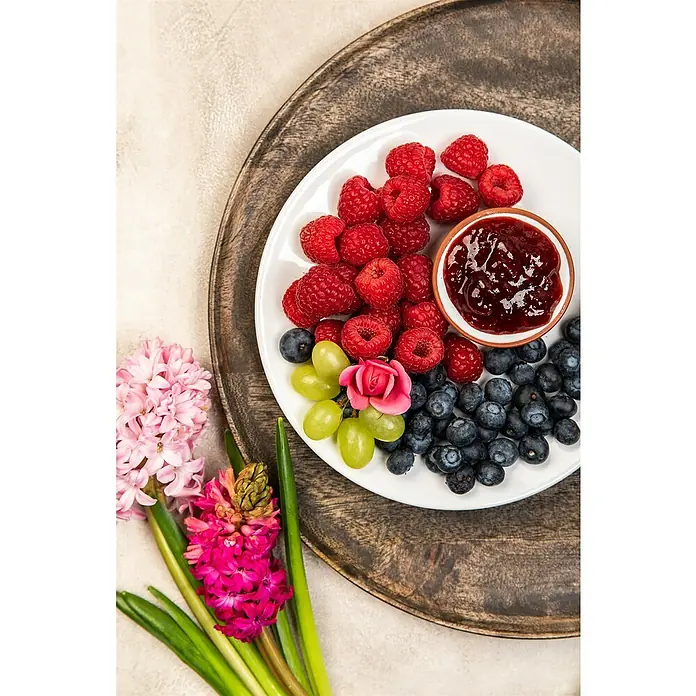 Piardino HimbeereRubus idaeus 'Sugana', Erntezeit: August - Festlicher Teller mit frischen Himbeeren, Blaubeeren, grünen Trauben und Konfitüre, dekoriert mit Blumen auf einem rustikalen Holztablett.