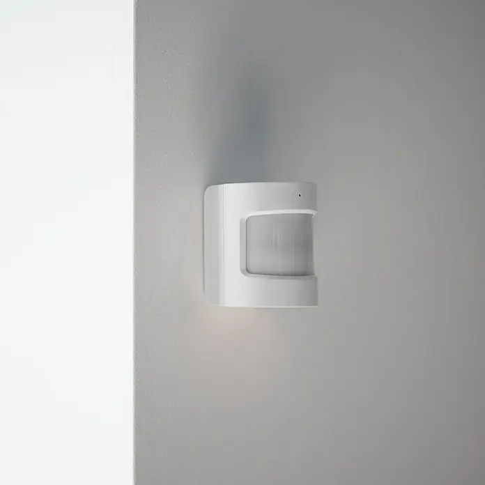 Hombli Bewegungsmelder - Reichweite Sensor: 8 m, Weiß, Erfassungswinkel Sensor: 120 °, L x B x H: 3,5 x 3,8 x 4,65 cm - Nahaufnahme eines weißen Bewegungsmelders an einer hellgrauen Wand, der Licht nach unten abstrahlt.