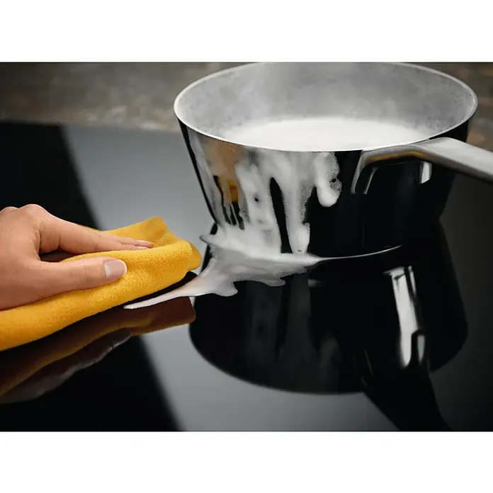 Electrolux Placa de cocción EIV83446 - Número de fogones de inducción: 4 ud. - Una olla con leche se derrama sobre una placa vitrocerámica que se está limpiando con un paño amarillo.