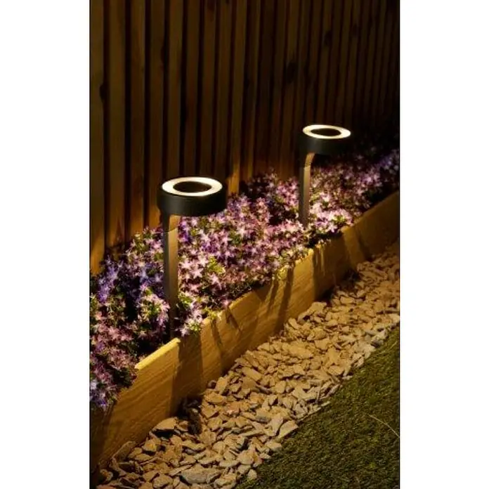 Estaca solar Orange - L x An x Al: 11 x 11 x 40 cm, Negro, IP33, 10 lm - Dos luces de jardín redondas iluminan flores y piedras junto a una valla de madera. Las luces son modernas y crean un ambiente acogedor.