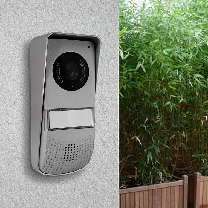 Avidsen Interfono con pantalla LCD - L x An x Al: 3,5 x 7 x 16,5 cm - La imagen muestra un timbre plateado con cámara montado en una pared, al lado de una jardinera de bambú exterior.