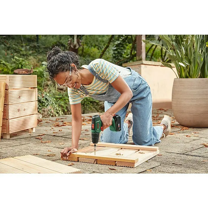 Bosch Power for All 18V Akku-Bohrschrauber EasyDrill 18V-40 - 18 V, 2 Akkus, 2 Ah, 40 Nm - Eine Frau kniet und benutzt einen grünen Bosch-Akkuschrauber, um ein Holzprojekt im Freien zu montieren.