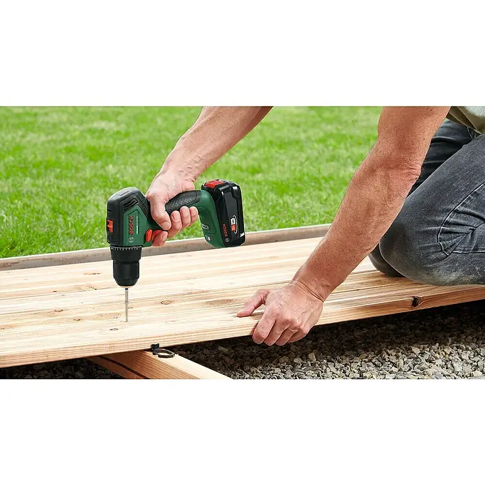 Bosch Power for All 18V Akku-Bohrschrauber UniversalDrill 18V-60 - 18 V, 2 Akkus, 60 Nm - Nahaufnahme eines Mannes, der mit einem Bosch Akku-Bohrschrauber ein Holzbrett auf einer Terrasse befestigt. Das grüne Gerät von Bosch ist deutlich sichtbar.