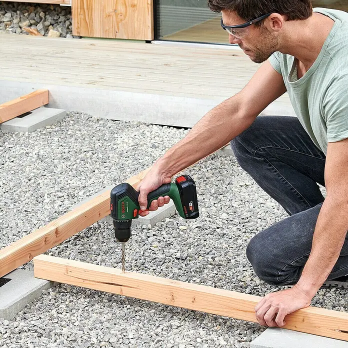 Bosch Power for All 18V Akku-Bohrschrauber UniversalDrill 18V-60 - 18 V, 2 Akkus, 60 Nm - Ein Mann arbeitet mit einem Bosch Akku-Bohrschrauber an einem Holzrahmen im Freien. Der grüne Bohrschrauber ist kompakt und leistungsstark.
