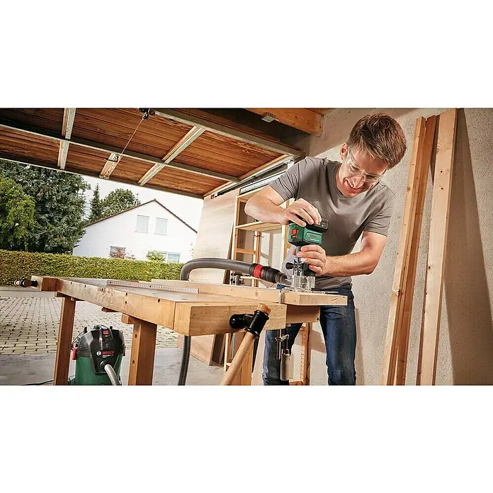 Bosch Power for All 18V Akku-Kombifräse AdvancedTrimRouter 18V-8 - 18 V, Ohne Akku, Leerlaufdrehzahl: 10.000 U/min - 30.000 U/min - Mann arbeitet mit einem Bosch-Elektrowerkzeug an einem Holztisch, mit Holzbrettern an der Seite und einem Staubsauger darunter.