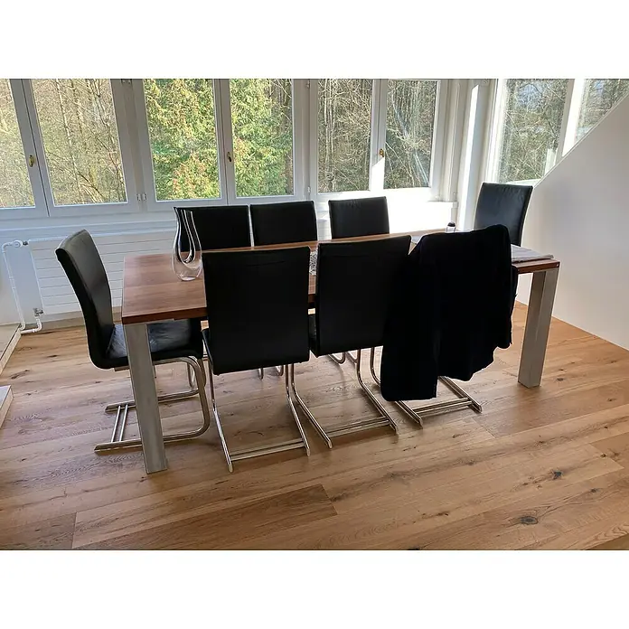 Parkett Eiche Wien - Eiche Wien, Landhausdiele, 1.250 x 190 x 14 mm - Esszimmer mit Esstisch, schwarzen Lederstühlen und einem transparenten Krug vor großen Fenstern mit Blick auf grüne Bäume.