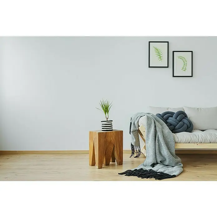 Erfurt Vliestapete Variovlies Flat Premium - Weiß, Uni, 25 x 0,75 m - Elegantes Interieur mit Holzhocker, Pflanze, Sofaecke mit Decke und Kissen sowie zwei gerahmten Pflanzenbildern an einer weißen Wand.