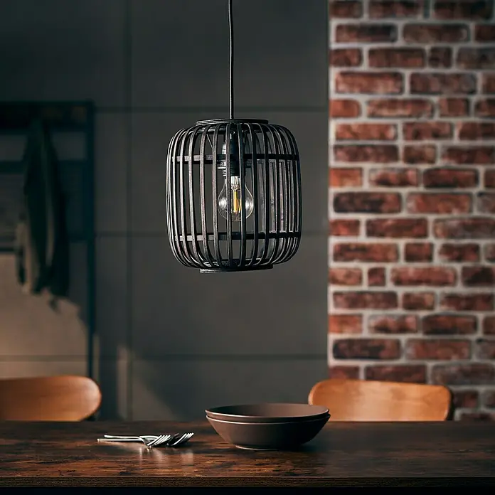 Brilliant Woodrow Pendelleuchte - E27, 60 W, Schwarz - Nahaufnahme einer dekorativen Pendelleuchte aus dunklem Metall mit einer Edison-Glühbirne über einem Holztisch.