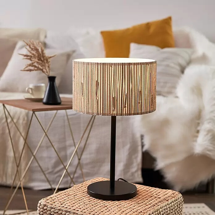 Brilliant Wimea Tischleuchte - 52 W, Schwarz, E27 - Tischlampe mit schwarzem Fuß und natürlichem Weblampenschirm, warmes Licht spendend. Sie steht auf einem Flechthocker neben einem Sofa.