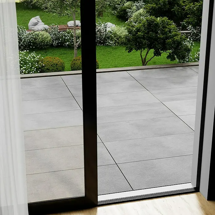 Cersanit Terrassenfliese Metropolis - 59,3 x 119,3 x 2 cm, Light Grey, Matt - Blick durch eine geöffnete Terrassentür auf eine gepflasterte Terrasse und einen Garten mit Rasen und Bäumen.