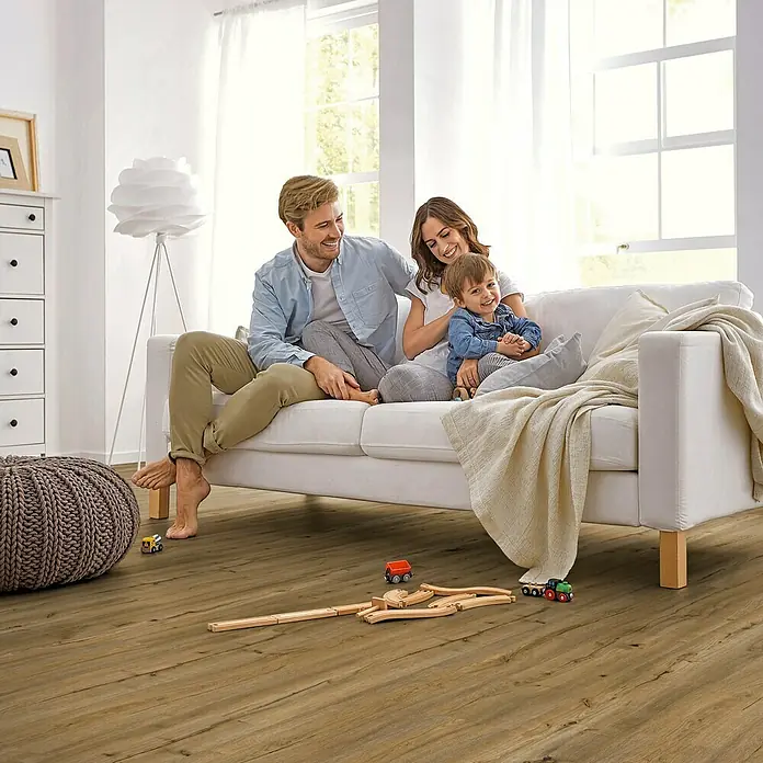 Decolife Vinylboden Comfort - Rustic Oak, Landhausdiele, 1.220 x 185 x 10,5 mm - Glückliche Familie entspannt auf dem Sofa in einem Wohnzimmer mit Holzboden und Spielzeug.
