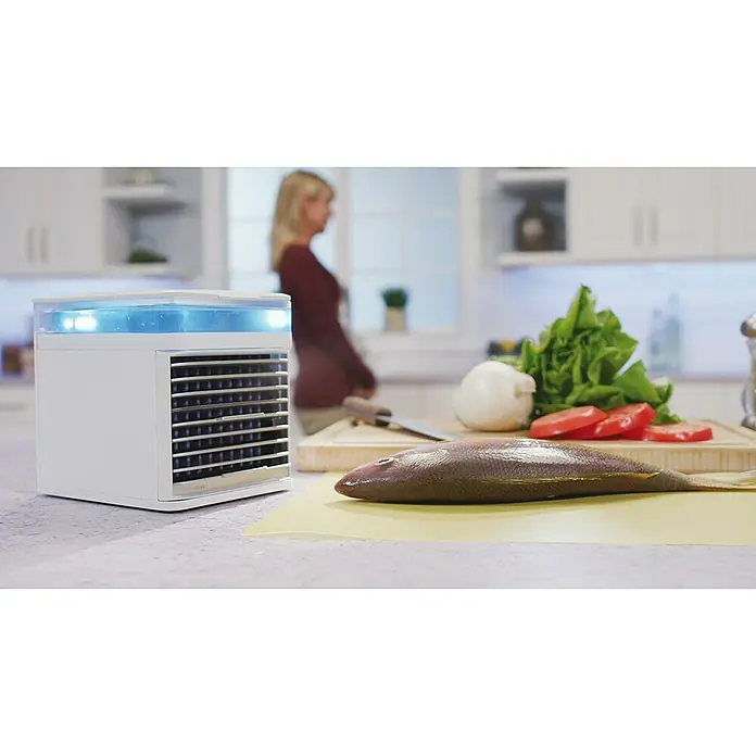 Arctic Air Climatizador evaporativo Pure Chill - L x An x Al: 17,8 x 20 x 24,8 cm - Un enfriador de aire compacto blanco con luz azul se sitúa en una encimera de cocina junto a ingredientes frescos.
