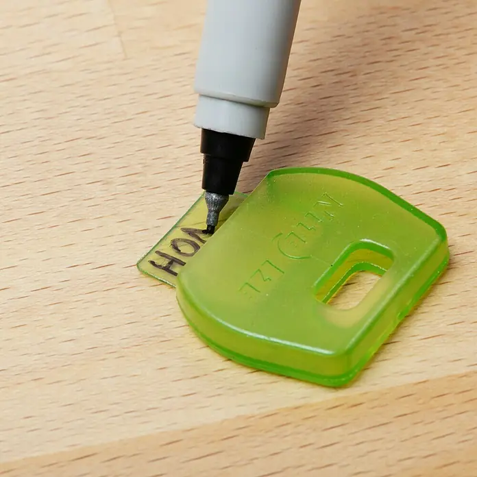 Nite Ize Schlüsselkappe - 4 Stk., Mehrfarbig - Nahaufnahme von einer transparent grünen Nulksen® Schlüsselkappe, die mit einem Stift beschriftet wird, auf einer Holzoberfläche.