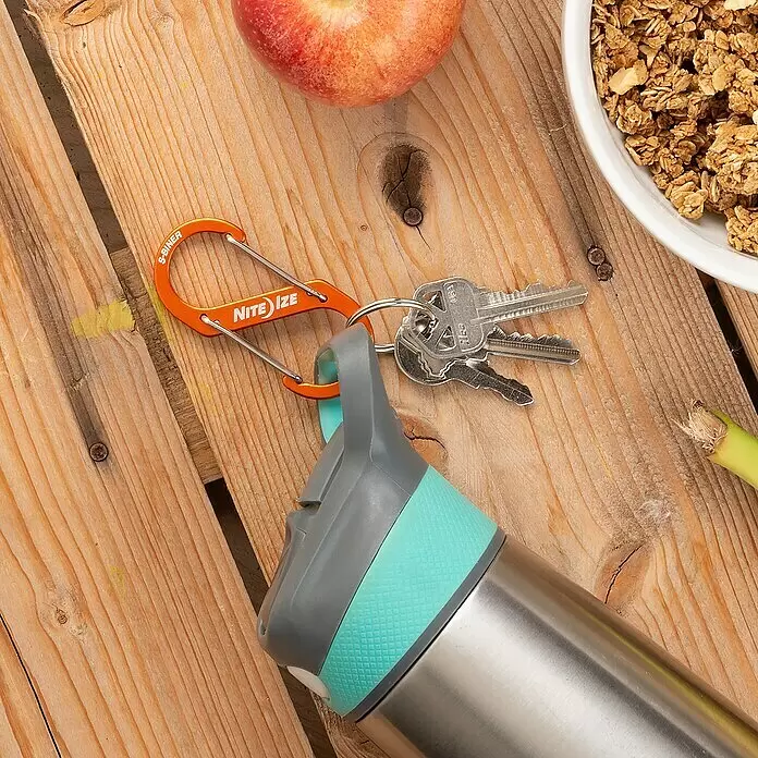 Nite Ize Mosquetón doble S-Biner - 69 x 31 mm - Mosquetón S-Biner naranja que sujeta llaves y se une a una botella de agua, sobre una mesa de madera con manzana y granola.
