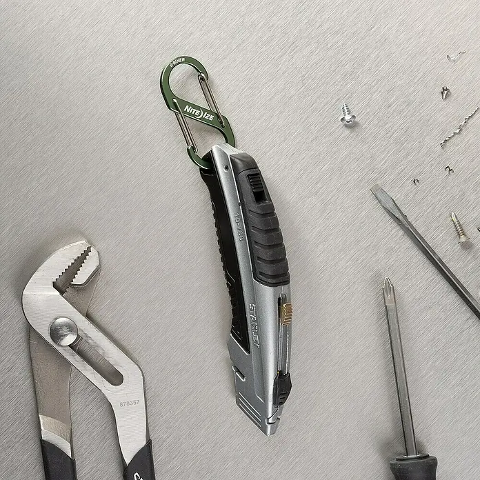 Doppelkarabiner S-Biner - Olivgrün - Stanley Cuttermesser mit Karabinerhaken, Schraubendreher und Rohrzange auf einer Werkbank.