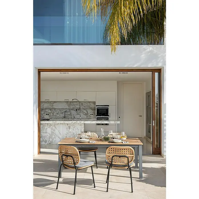 Sunfun Carina Gartentisch - L x B x H: 180 x 100 x 75 cm, Akazie, Stahl, Rechteckig, Hellbraun - Bild eines Esstisches mit Rattanstühlen auf einer Terrasse, die einen Blick in eine moderne Küche mit Marmorinsel bietet.
