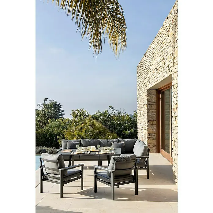 Conjunto de muebles Simona - 7 pzs., Acero/Polywood, Gris oscuro - Un elegante conjunto de comedor exterior con cojines grises y una mesa puesta, con un muro de piedra de fondo. Diseño elegante para jardines y terrazas.