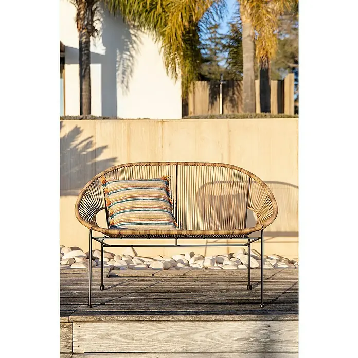 Sunfun Sillón de jardín Fidji - Marrón - Cómodo sofá de mimbre con un cojín colorido en una terraza de madera, con una pared brillante y palmeras al fondo.