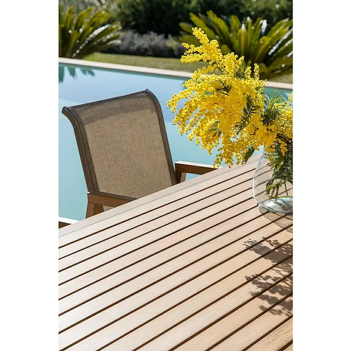 Conjunto de muebles Flavium - 7 pzs., Textiline/Enviro Imitation Wood®, Natural - Una imagen de una mesa con una silla al lado. Sobre la mesa, hay un jarrón con flores amarillas. Al fondo, hay una piscina.