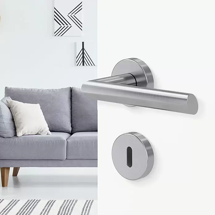 Diamond Doors Modern Drückergarnitur South Dakota - Stärke Tür: 40 mm - 45 mm, Edelstahl, L-Form, Buntbart - Türdrücker und passendes Schlüssellochschild aus gebürstetem Edelstahl an weißer Tür, mit modernem Wohnzimmer im Hintergrund.