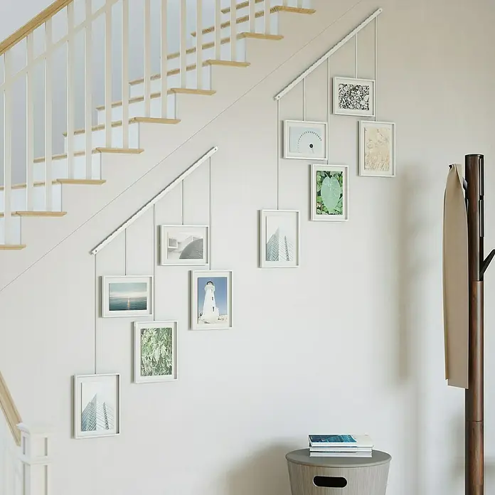Umbra Bilderrahmen-Set Exhibit - 2 x 10x15 cm, 3 x 13x18 cm, Weiß, Metall - Bilderrahmen an der Wand entlang einer Treppe befestigt. Die Rahmen sind weiß und enthalten verschiedene Motive wie eine Küstenlandschaft.