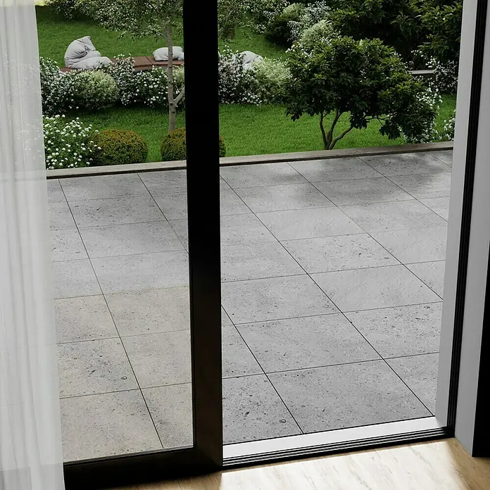 Marazzi Terrassenfliese Esterno 20T Gris Fleury - 60 x 60 x 2 cm, Grau, Matt - Blick durch eine Schiebetür auf eine gepflasterte Terrasse und einen Garten. Die Tür ist dunkel gerahmt und gibt den Blick frei.