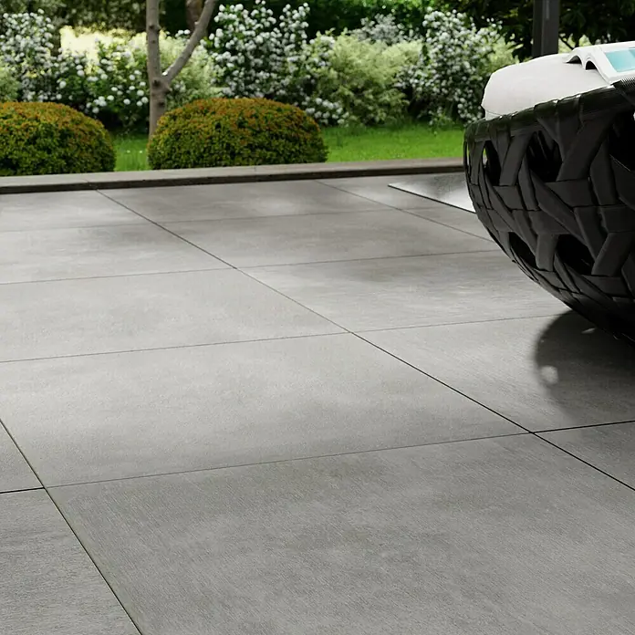 Marazzi Terrassenfliese Esterno 20T Plaster Grey - 60 x 60 x 2 cm, Grau, Matt - Nahaufnahme von grauen Terrassenfliesen mit einer geflochtenen schwarzen Sitzgruppe und Garten im Hintergrund.