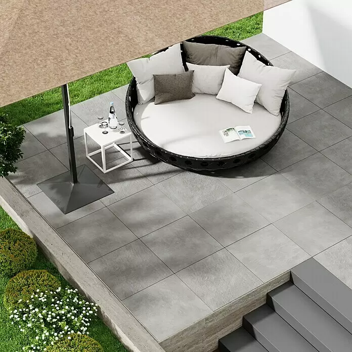 Marazzi Terrassenfliese Esterno 20T Plaster Grey - 60 x 60 x 2 cm, Grau, Matt - Rundes schwarzes Rattan-Outdoor-Tagesbett mit hellgrauen Polstern auf einer Terrasse unter einem großen beigefarbenen Sonnenschirm.