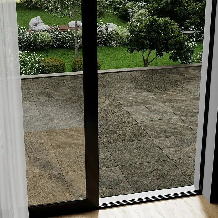 Marazzi Terrassenfliese Esterno 20T Rocking Grey - 60 x 60 x 2 cm, Grau/Braun, Matt - Blick durch eine Terrassentür auf eine Steinterrasse, die in einen gepflegten Garten übergeht. Die Fliesen sind quadratisch und haben eine raue, natürliche Oberfläche.