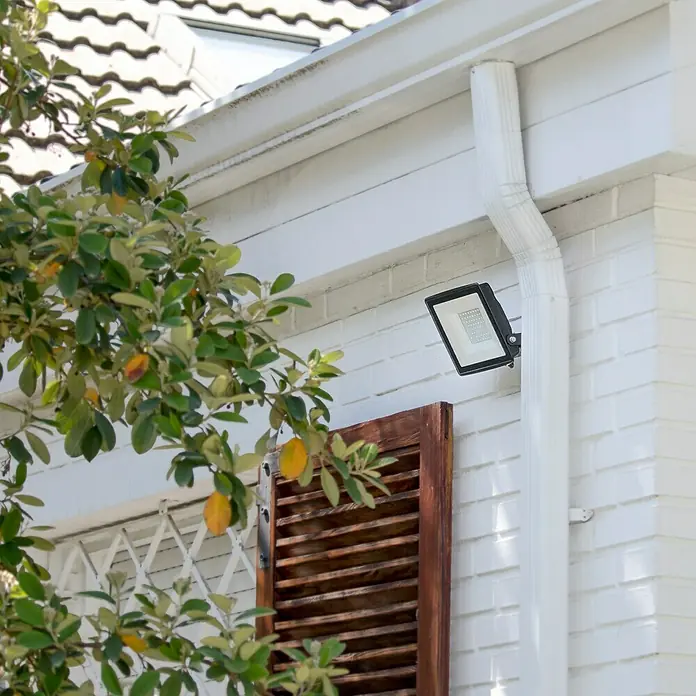 Garza Proyector LED Basic - 10 W - Moderna lámpara LED exterior en una pared de ladrillo blanco con canaleta y ventana parcialmente oculta por un árbol.
