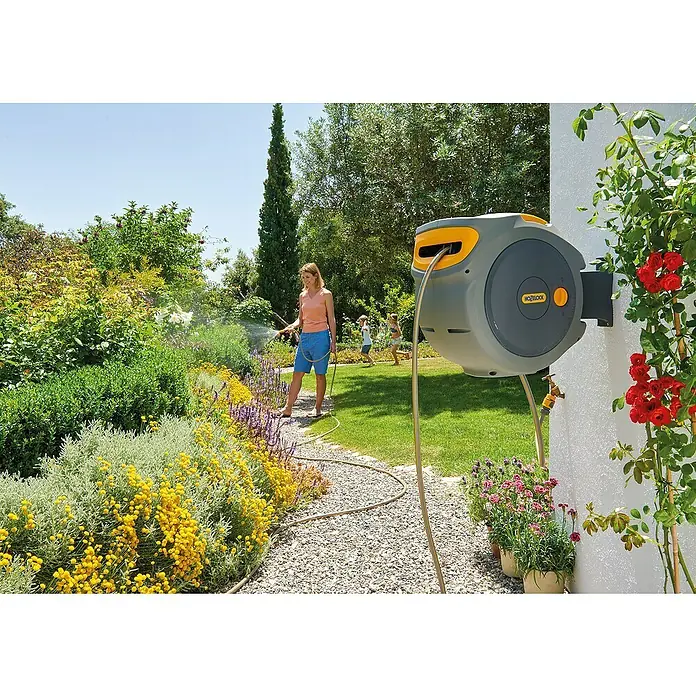 Hozelock Wand-Schlauchbox Auto Reel - Schlauchlänge: 20 m - Frau benutzt eine Hozelock Schlauchbox zum Bewässern von Pflanzen in einem sonnigen Garten, während Kinder im Hintergrund spielen.