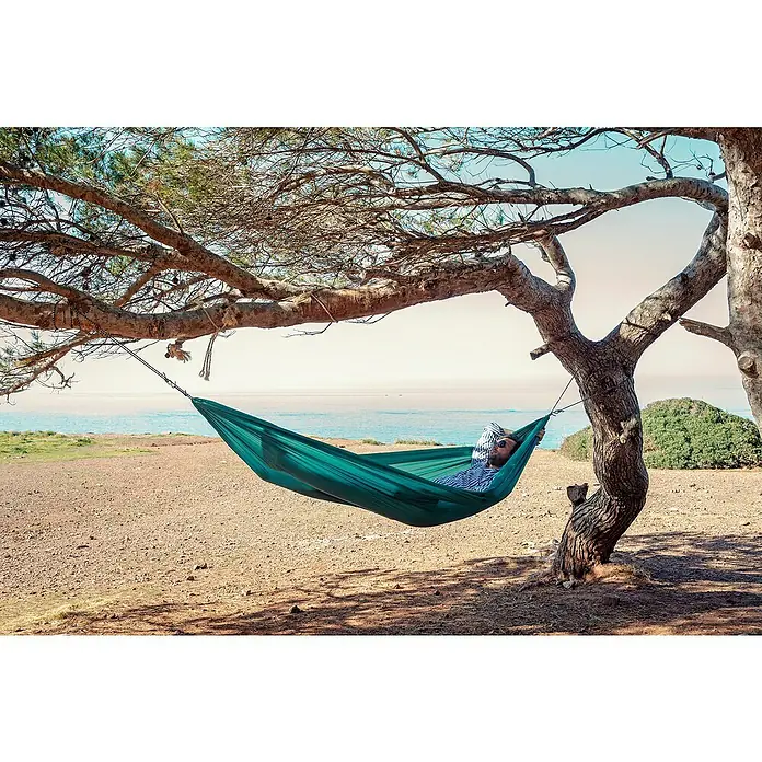 Amazonas Hängematte Travel Set - L x B: 275 x 140 cm, Grün - Entspannung pur: Ein Mann ruht sich in einer grünen Hängematte zwischen Bäumen am Strand aus und genießt die Aussicht auf das Meer.