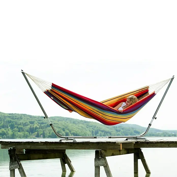 Amazonas Hängematte Barbados - Rainbow - Ein farbenfrohes Hängemattenset mit Gestell, perfekt zum Entspannen im Freien. Die Hängematte ist auf einem Holzsteg am See aufgebaut.