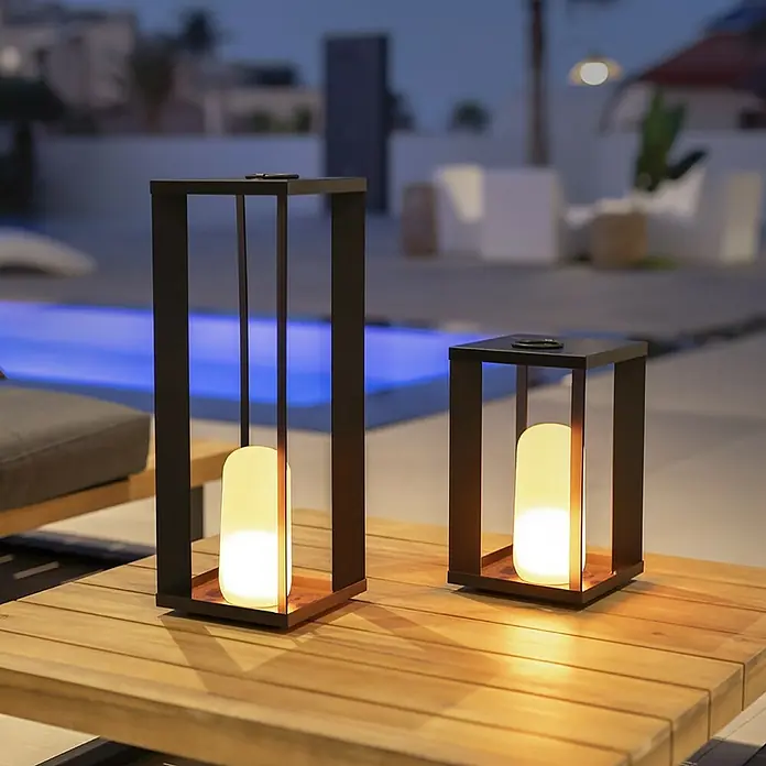 New Garden Außenlaterne -  - Zwei dekorative, moderne Laternen mit warmem Licht auf einem Holztisch im Freien, die eine gemütliche Atmosphäre schaffen.