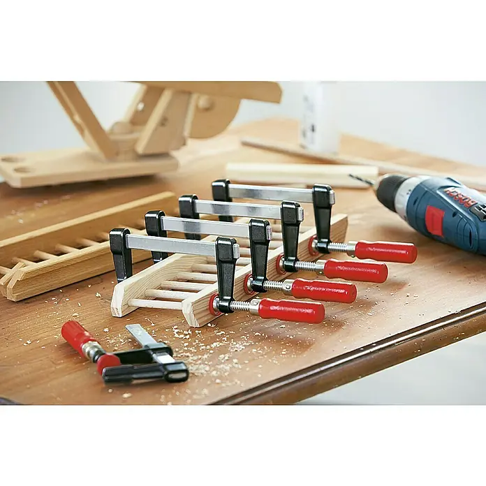 Bessey Schraubzwinge LM 250/50 - Spannweite: 250 mm, Spannkraft: 2.000 N, Ausladung: 50 mm - Nahaufnahme von Tischklemmen, die ein Holzprojekt sichern, zusammen mit einem Akkuschrauber auf einem Holztisch.