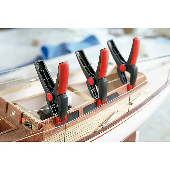 Bessey Federzwinge Clippix XC 2 - Spannweite: 25 mm, Spannkraft: 40 N, Ausladung: 30 mm - Drei Federklemmen befestigen die Holzverkleidung eines Schiffsmodells, wobei das rote und schwarze Design der Klemmen hervorsticht.