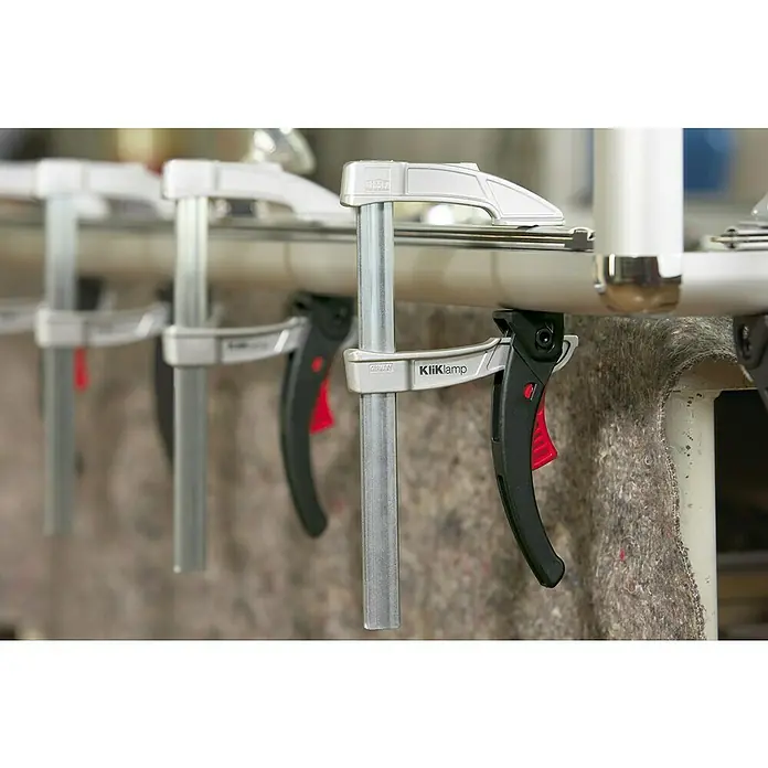 Bessey Hebelzwinge KliKlamp KLI 16 - Spannweite: 160 mm, Spannkraft: 1.200 N, Ausladung: 80 mm - Mehrere KliKlamp-Schnellspannhebelzwingen halten ein Rohr fest. Die schwarzen Griffe und roten Knöpfe stechen hervor. Professionelles Werkzeug.